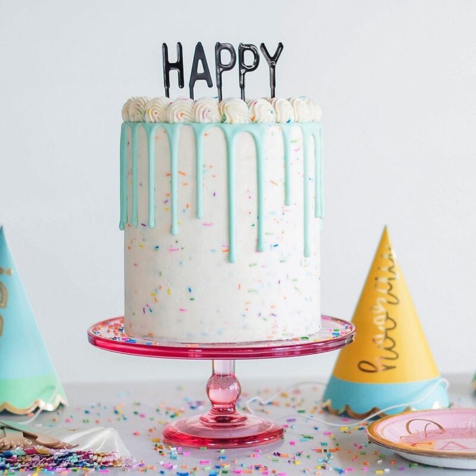 A tall cake with white frosting and blue drips sits on a pink stand. The cake is topped with the word "HAPPY" and decorated with colorful sprinkles. Nearby are party hats and confetti on the table.