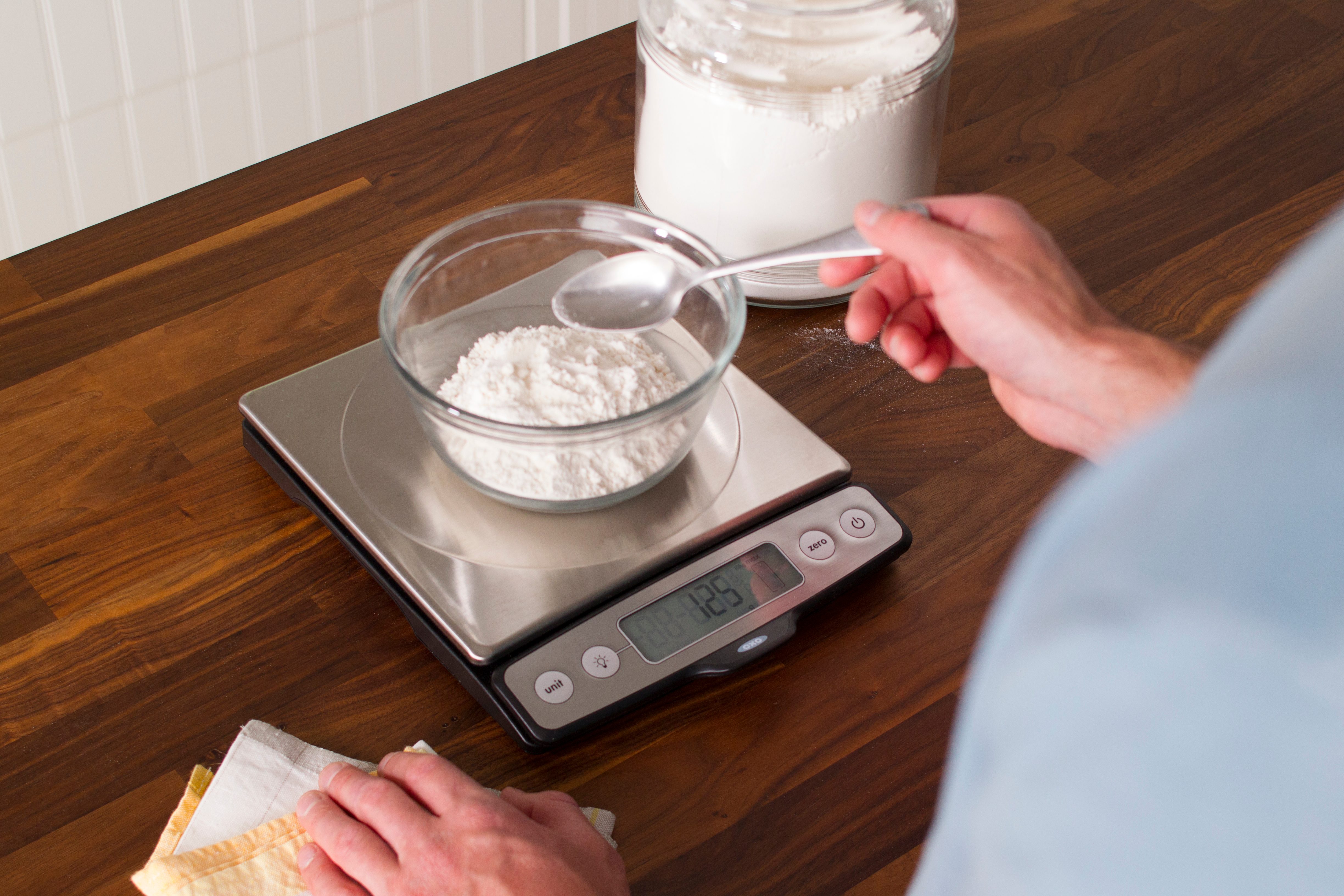 Person using a spoon to scoop flour into a small bowl on a scale