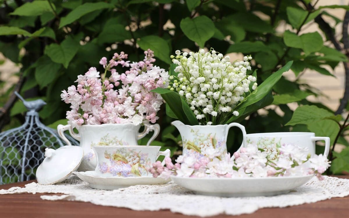 Lilac, bunch, bouquet of lily of the valley in vintage, antique authentic china, porcelain milk jug, sugar bowl, tea cup with saucer on lace napkin on table, bush background, garden floral scene