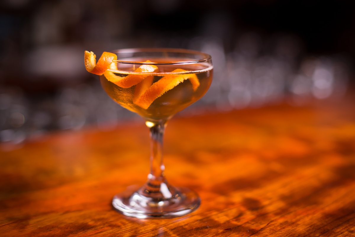 Low angle closeup of gourmet old fashion martini drink in champagne saucer cocktail glass filled with bourbon and brandy garnished by delicately spiral orange peel on red cherry wood grain countertop.
