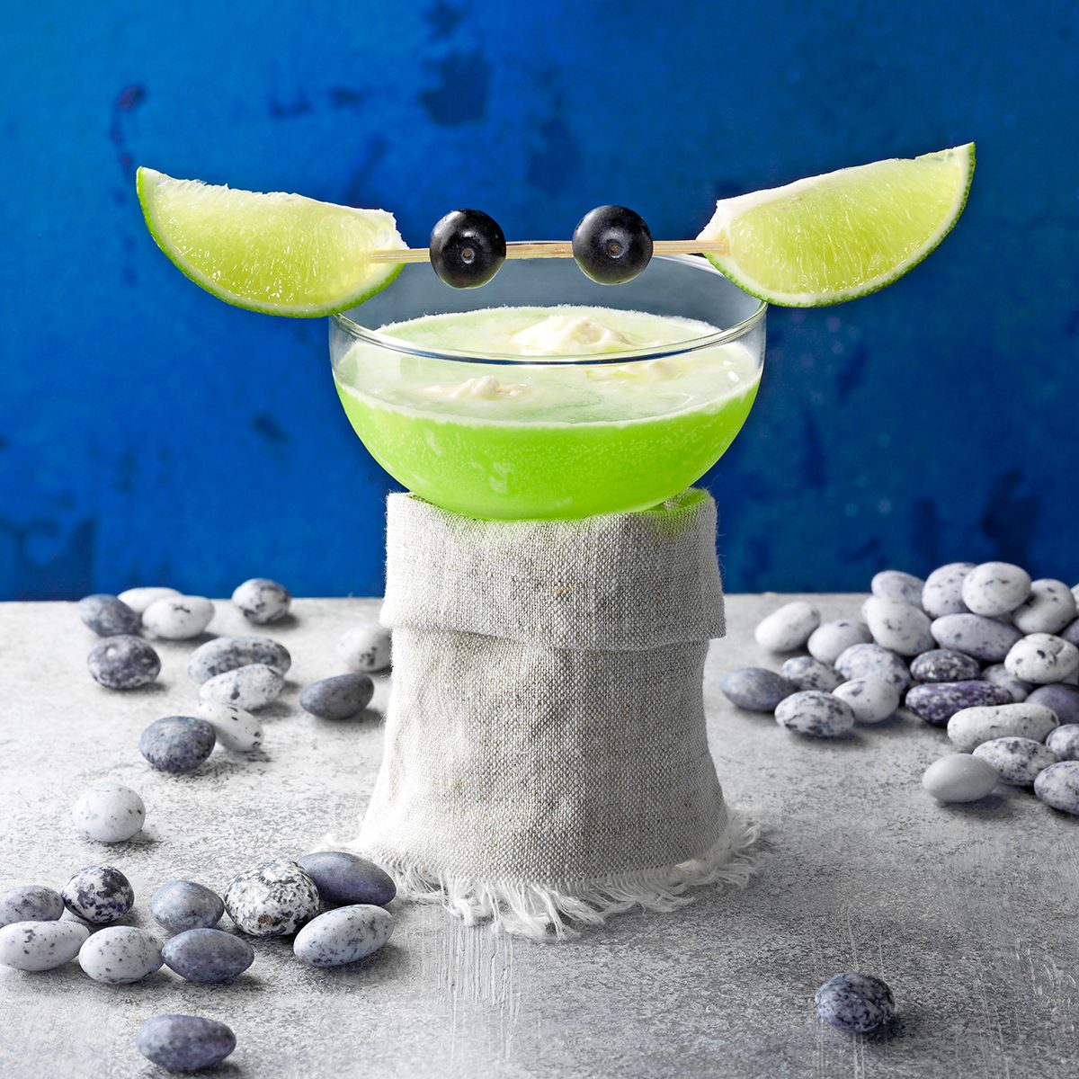 Yoda Soda; grey surface; gray surface; blue background; copy space; low camera angle; star wars; rock candy; blueberries; limes; lime wedges; grey; cloth; gray cloth; fantasy