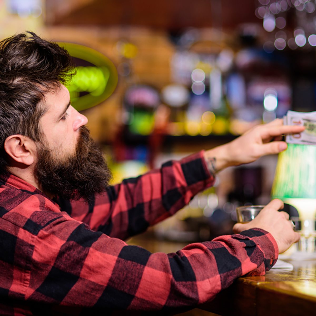 Man with drunk face sit alone at bar counter. Hipster holds glass with alcoholic drink and money, ordering more drinks.