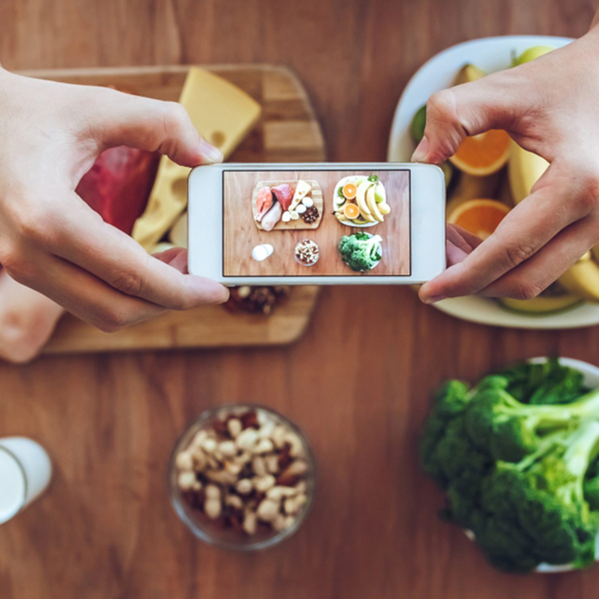 Top view of meat, fish, fruits, vegetables, nuts, milk are lying on the table while man is taking photo of it on a smart phone.
