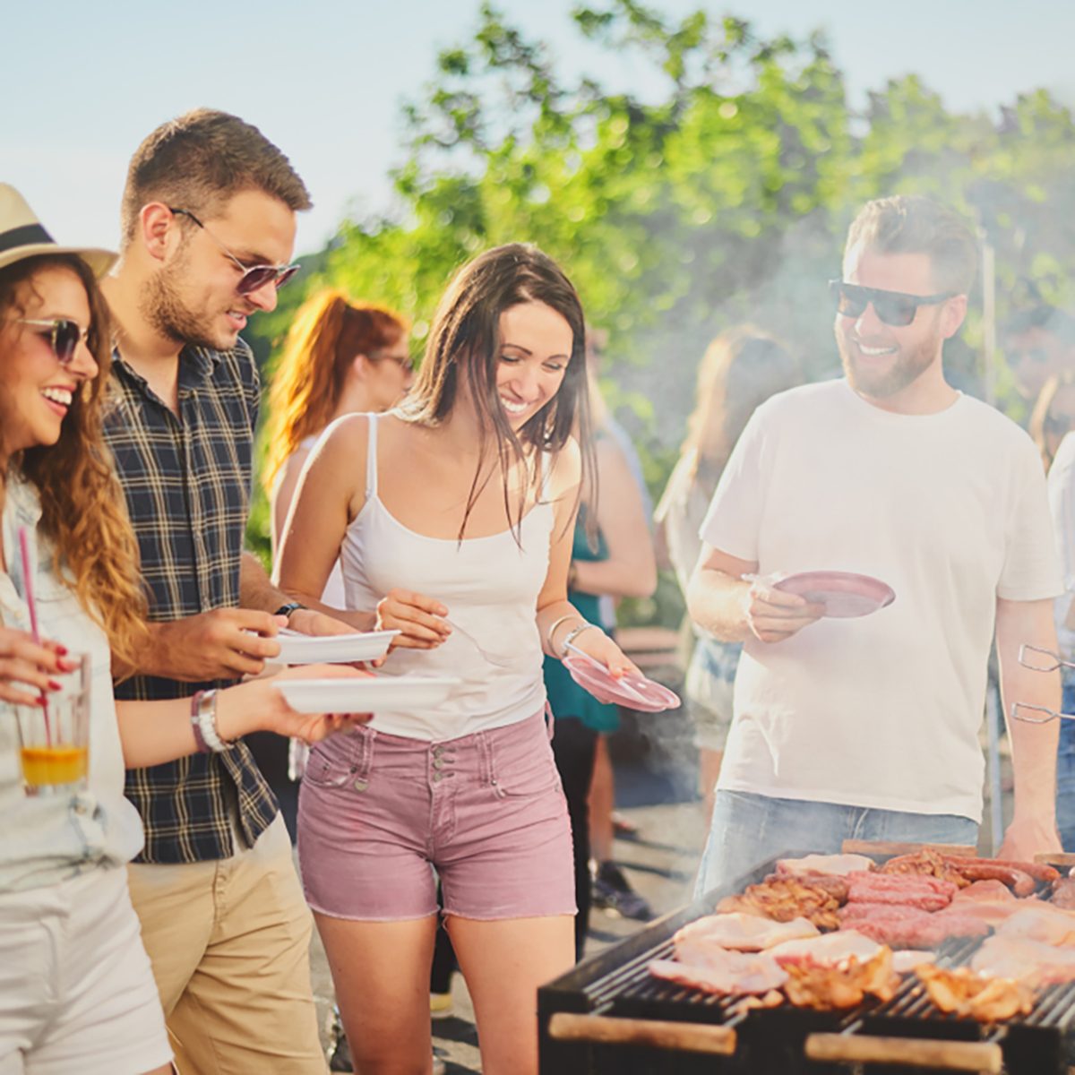 Group of people standing around grill, chatting, drinking and eating. 