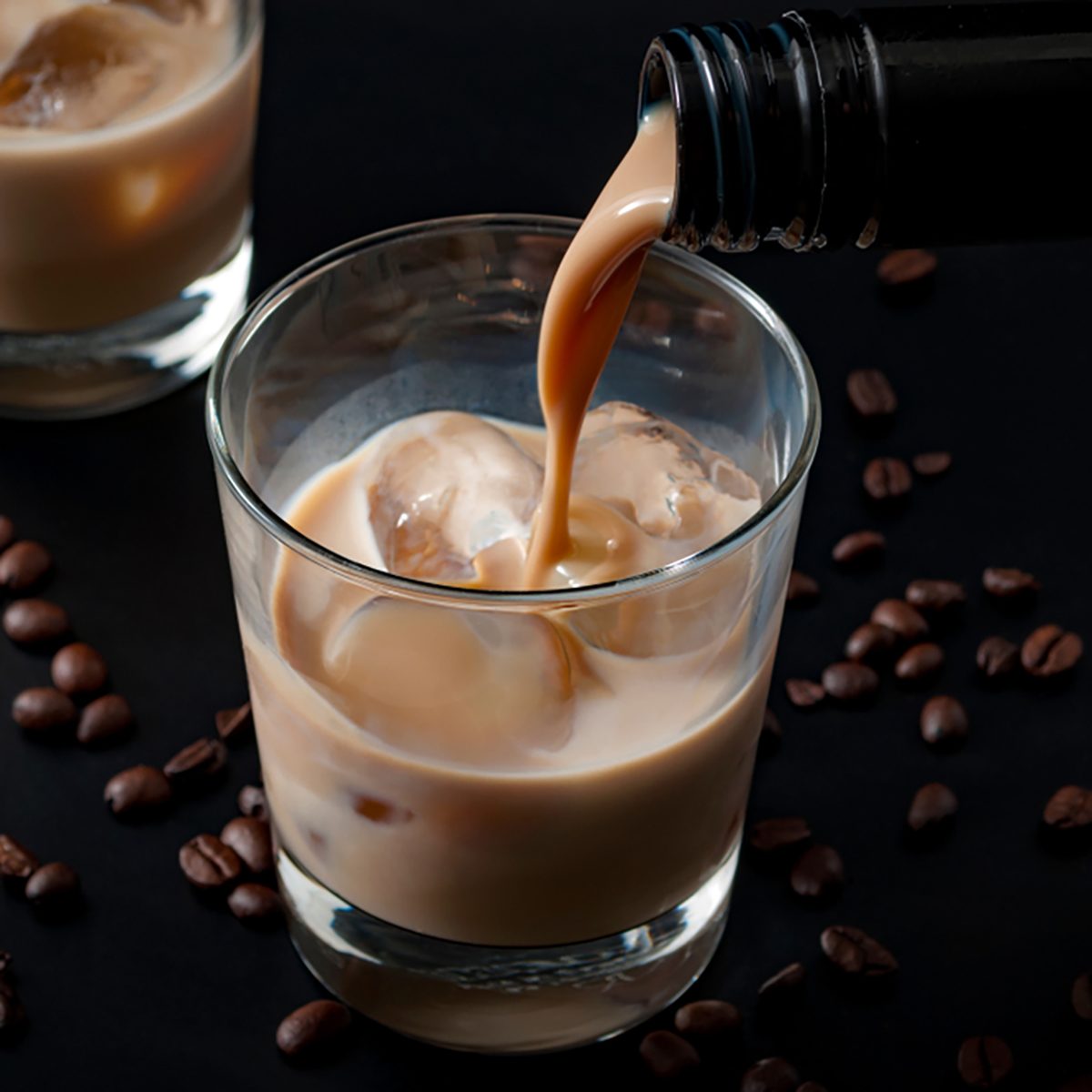 Pouring irish cream in a glass with ice, surrounded by coffee beans on a dark black background