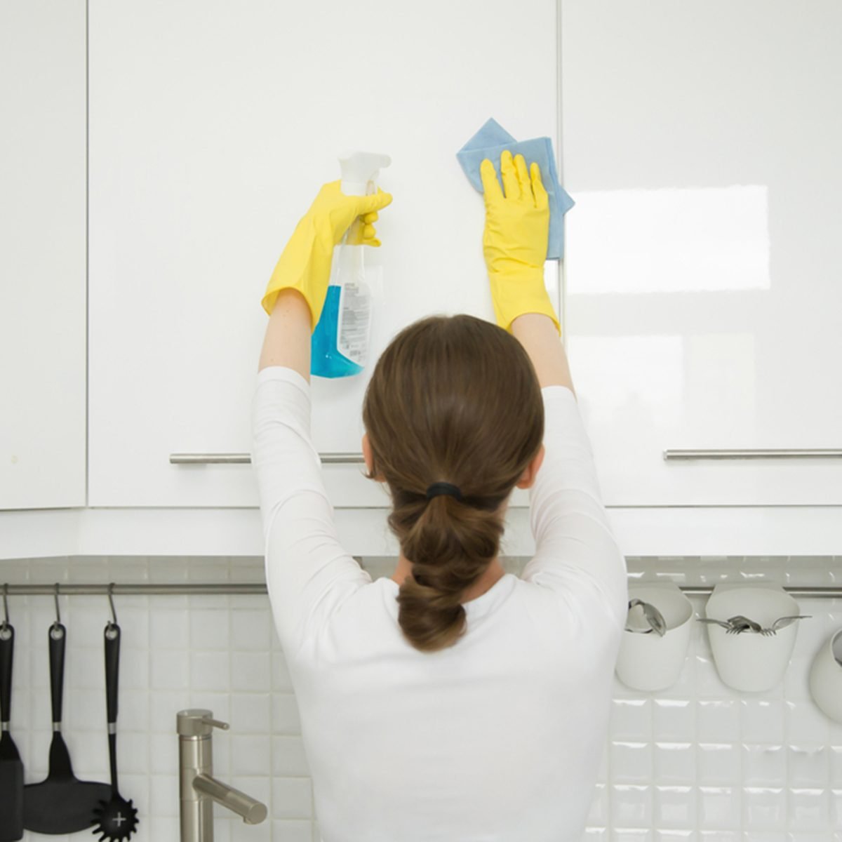 Rear view at an attractive young woman cleaning a surface of white kitchen wall cabinet, wearing rubber protective yellow gloves, with rag and spray bottle detergent.