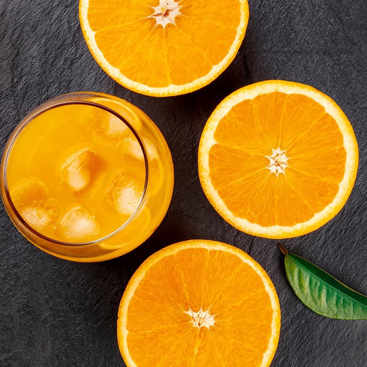 A closeup photo of a glass of fresh orange juice with ice cubes, orange halves and a green leaf, shot from the top on a black background
