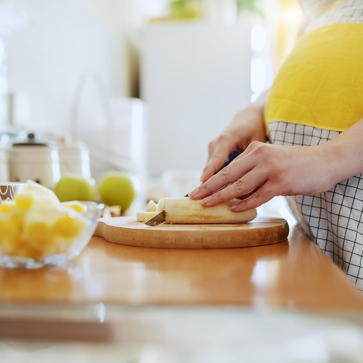 Close up of Caucasian pregnant woman in apron cutting banana and preparing healthy breakfast.