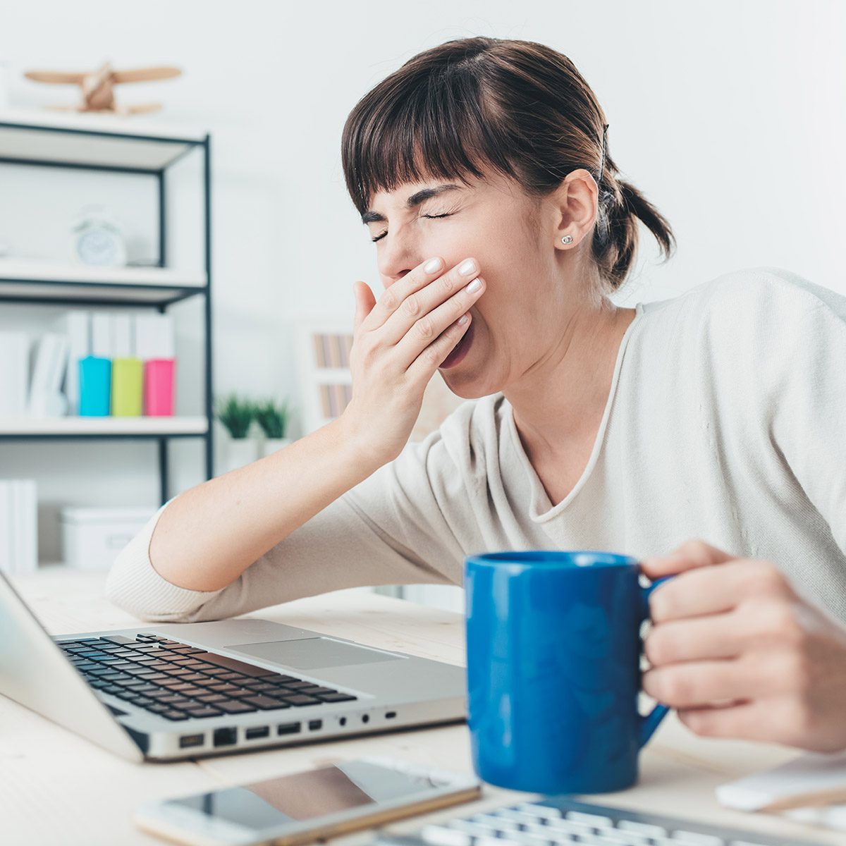 Tired sleepy woman yawning, working at office desk and holding a cup of coffee, overwork and sleep deprivation concept; Shutterstock ID 367741853