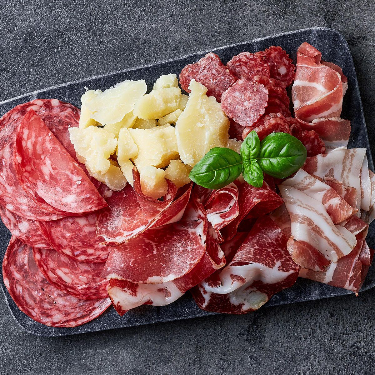 Marble cutting board with prosciutto, bacon, salami and cheese on dark stone background.