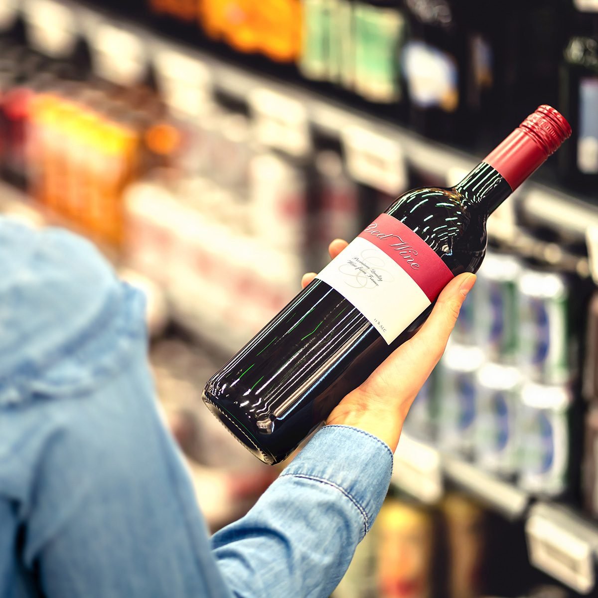 Woman reading the label of red wine bottle in liquor store or alcohol section of supermarket.