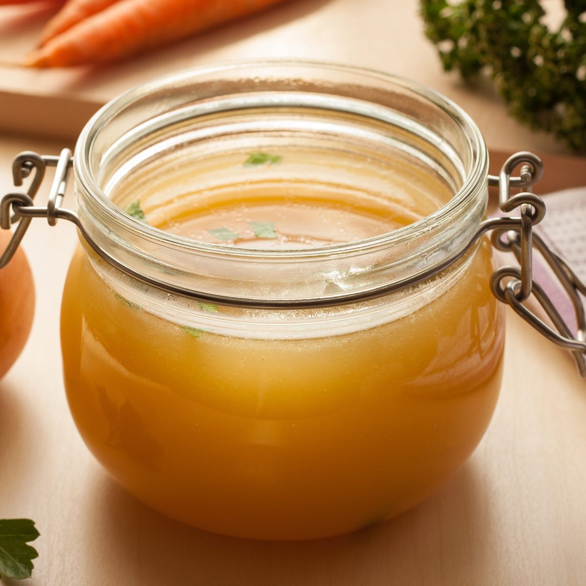 Bone broth made from chicken in a glass jar, with carrots, onions, and parsley in the background