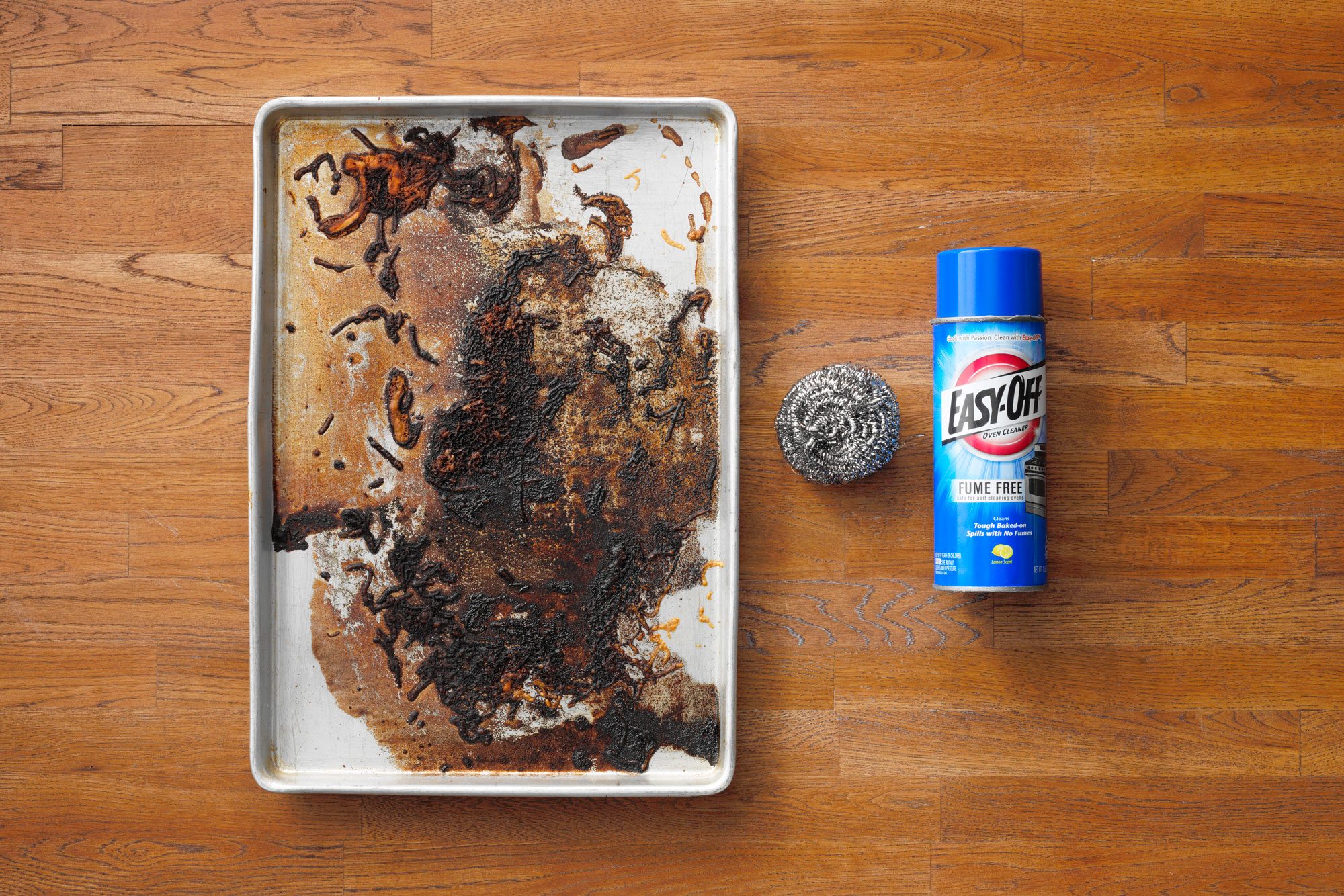 Dirty Baking Sheet next to steel wool and Easy Off Oven Cleaner on wood butcher block background