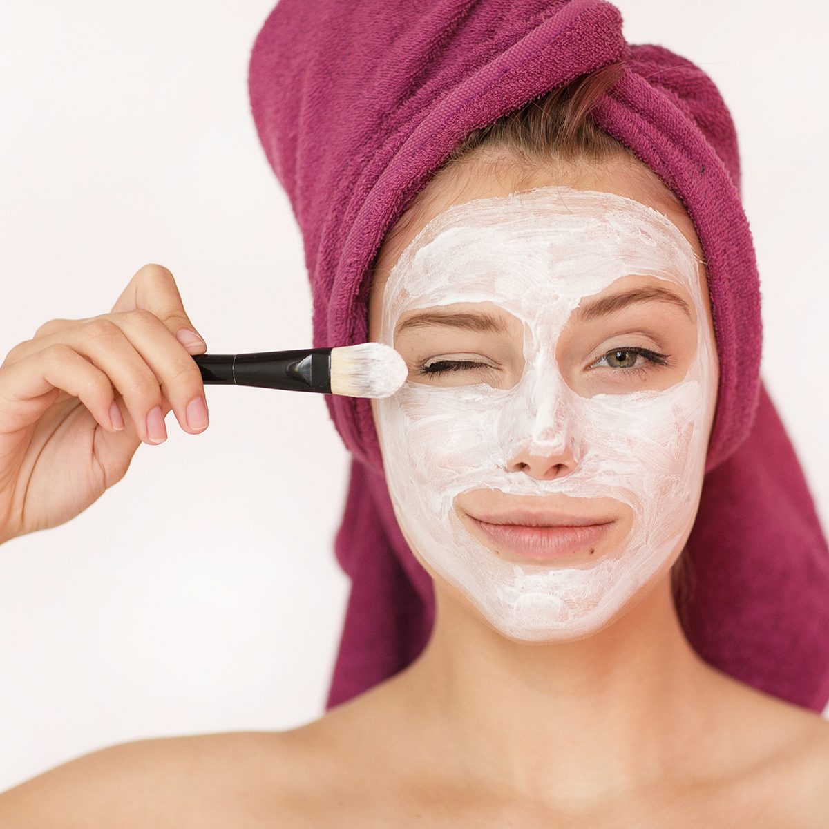 Happy and cheerful beautiful woman with a towel on her head and a cosmetic mask of white clay on her face looking into the camera applying a brush solution on her cheek isolated on a white background