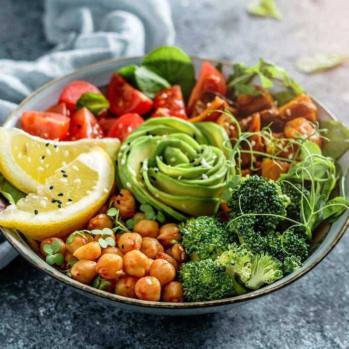 Buddha bowl salad with baked sweet potatoes, chickpeas, broccoli, tomatoes, greens, avocado, pea sprouts on light blue background with napkin.