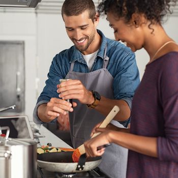 Young man cooking with girlfriend and adding spice to the sauce. Guy adds black pepper into frying pan on stove while woman using spatula to mix. Multiethnic couple preparing lunch together at home.