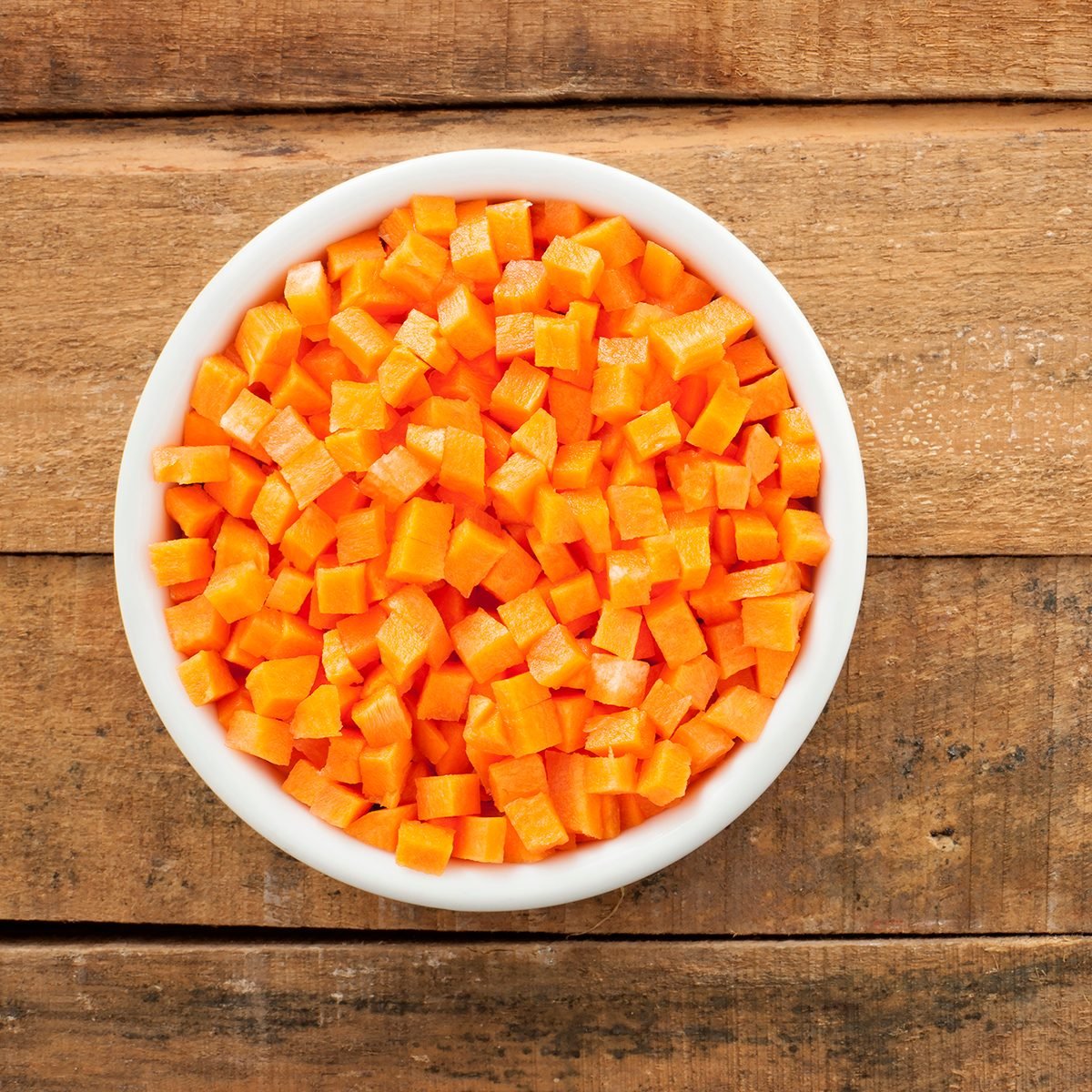 Top view of white bowl full of small carrot cubes over wooden table