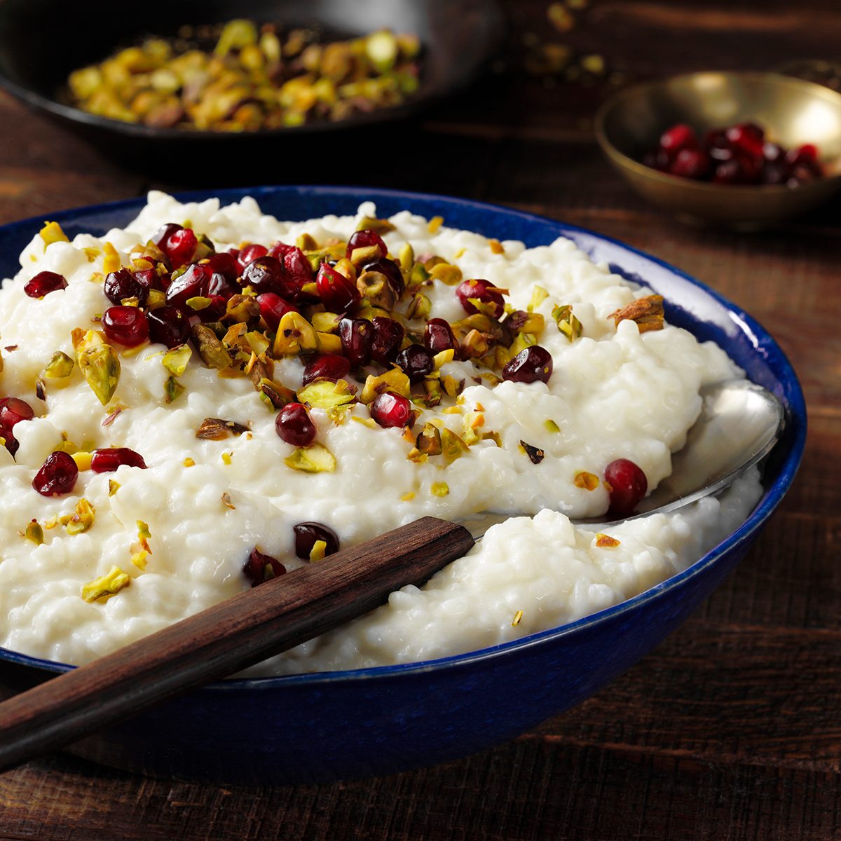 GLOBAL FLAVORS GO MAINSTREAM​ Food Trends Report, Rose Water Rice Pudding; 3/4 camera angle; wood surface; dark wood surface; russian wood surface; blue bowl; rose water; dark bowls; long grain rice; half-and-half cream; rose water; Pomegranate seeds; chopped pistachios; wood handled spoon; spoon; serving spoon