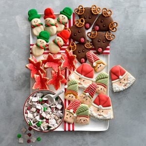 A platter of festive holiday cookies shaped like snowmen, reindeer, stars, elves, and Santa, with a bowl of peppermint candy mix; all are decorated with colorful icing and sprinkles on a gray surface.