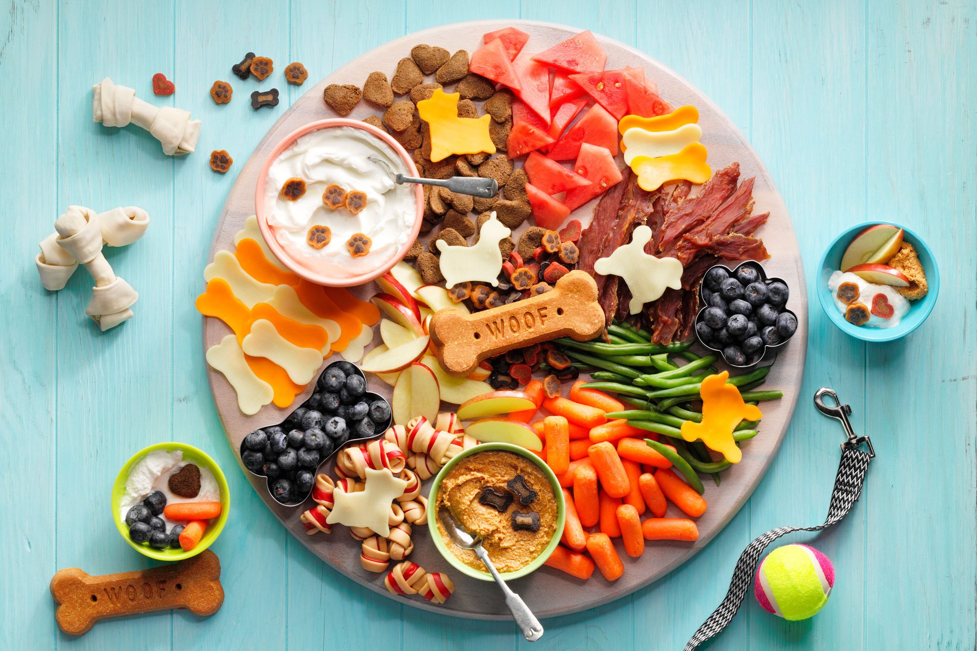 close up of "Barkuterie" dog charcuterie Board on light blue wood surface with dogs leash, toys, and treats around