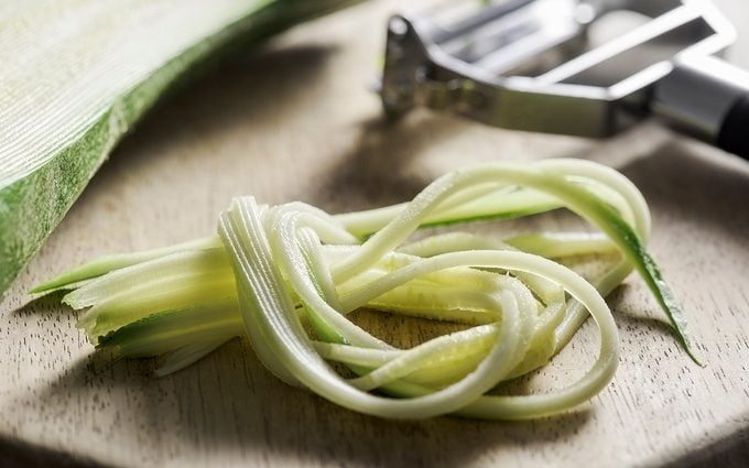 how to grate zucchini Strands of a julienned courgette are lying at the front of a wooden chopping board. The Julienne utensil and the rest of the courgette are lying behind it. The peeler is made from stainless steel and has a black handle. The selective focus is on the courgette in the foreground.