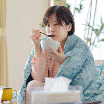 Sick Woman wrapped in a blanket eating soup on the couch
