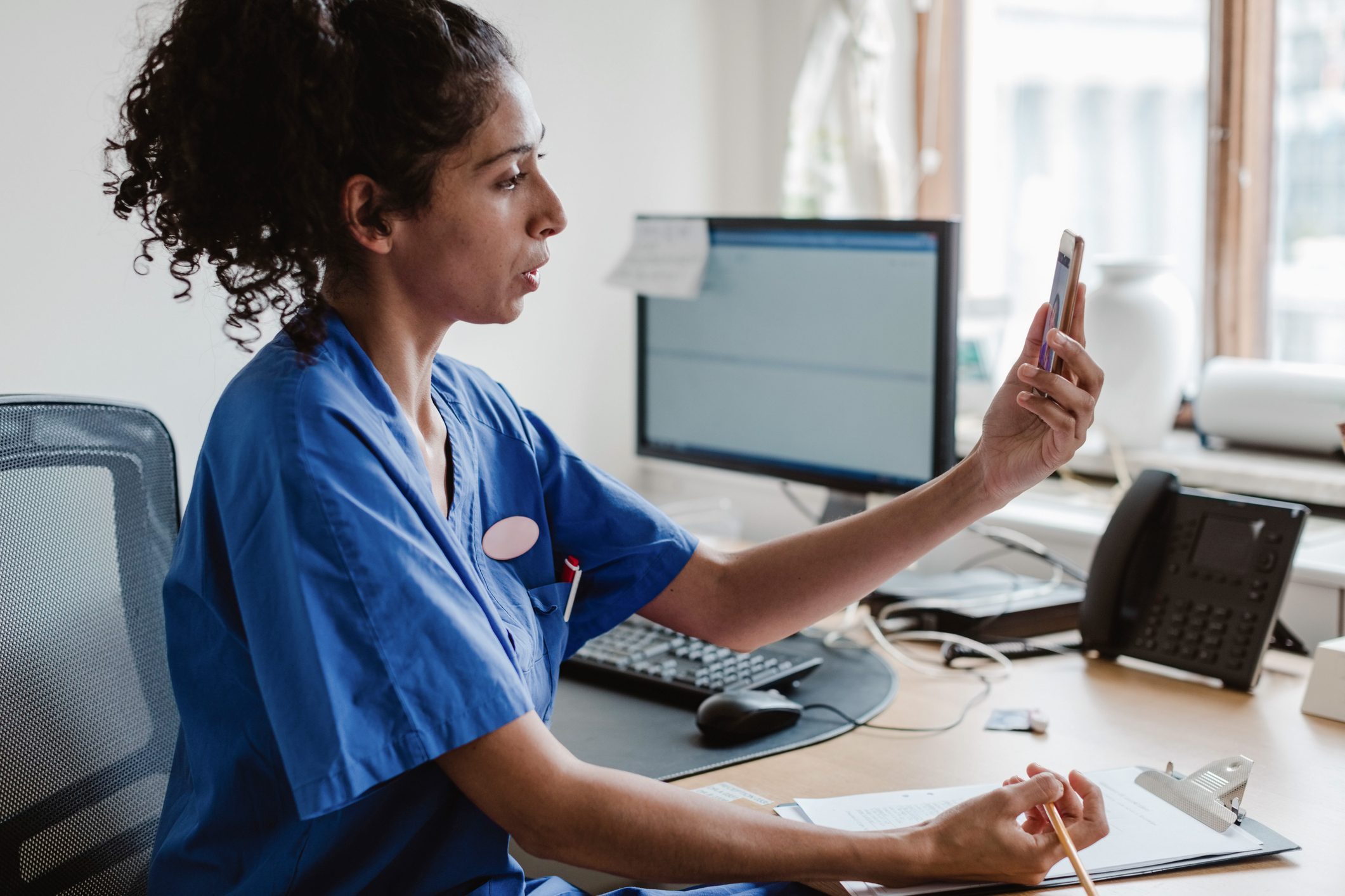 Female Healthcare Worker Talking On Video Conference Through Mobile Phone In Doctor