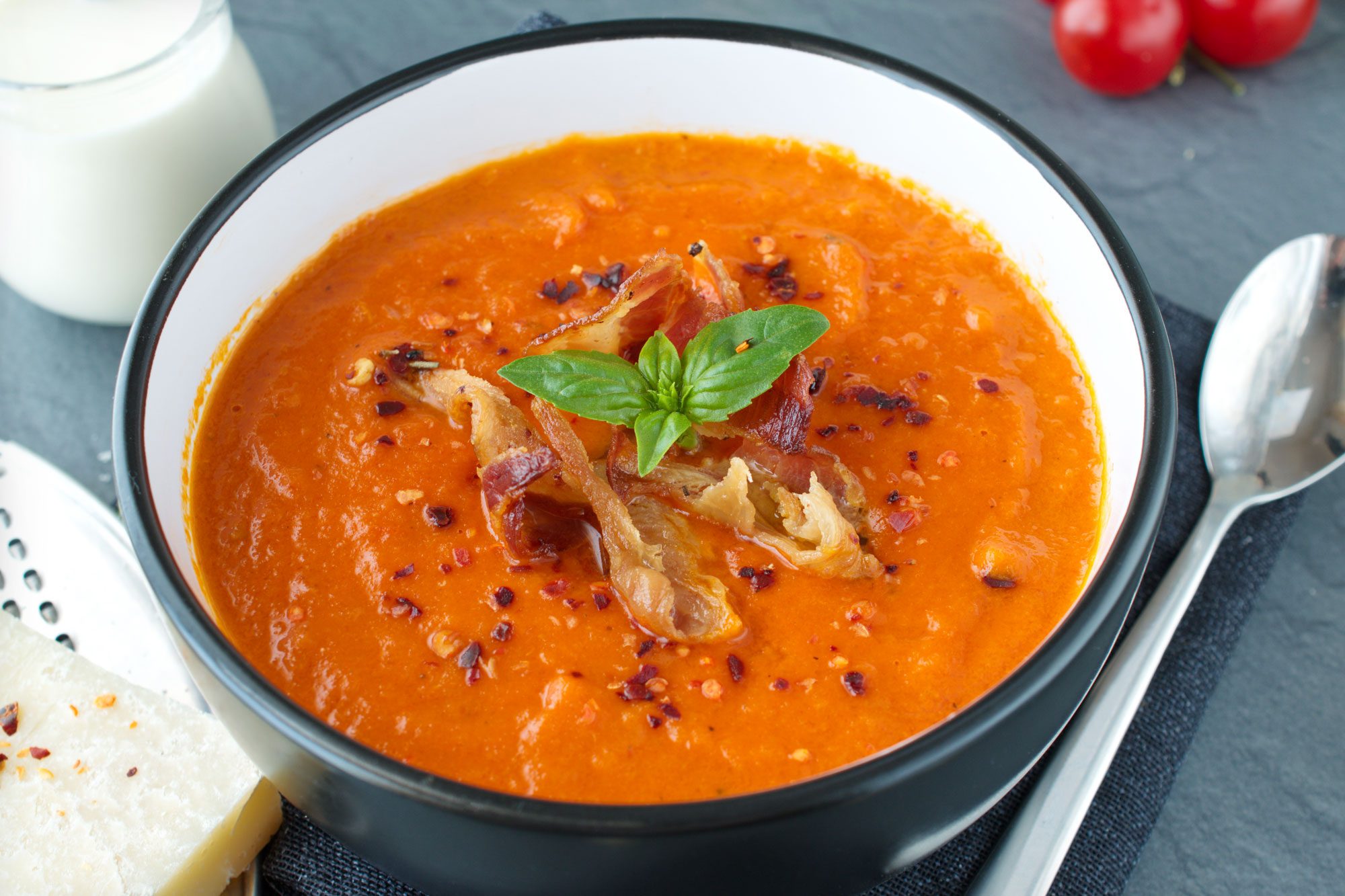 Tomato Soup With Basil And Fried Bacon In A Black Ceramic Bowl On A Grey Abstract Background