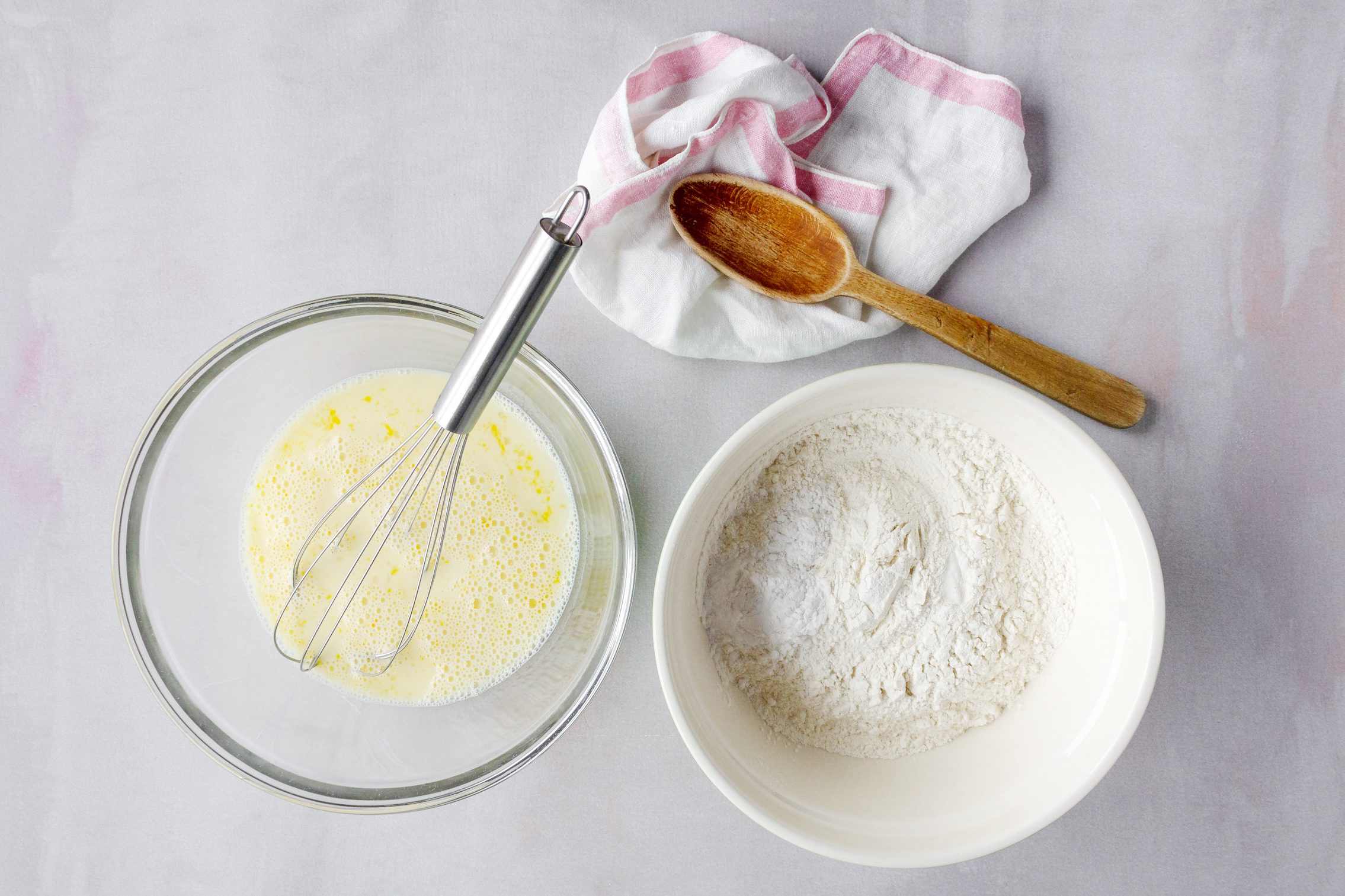 two bowls size by side on a counter, one with wet ingredients and a whisk and the other with dry ingredients and a wooden spoon on the side