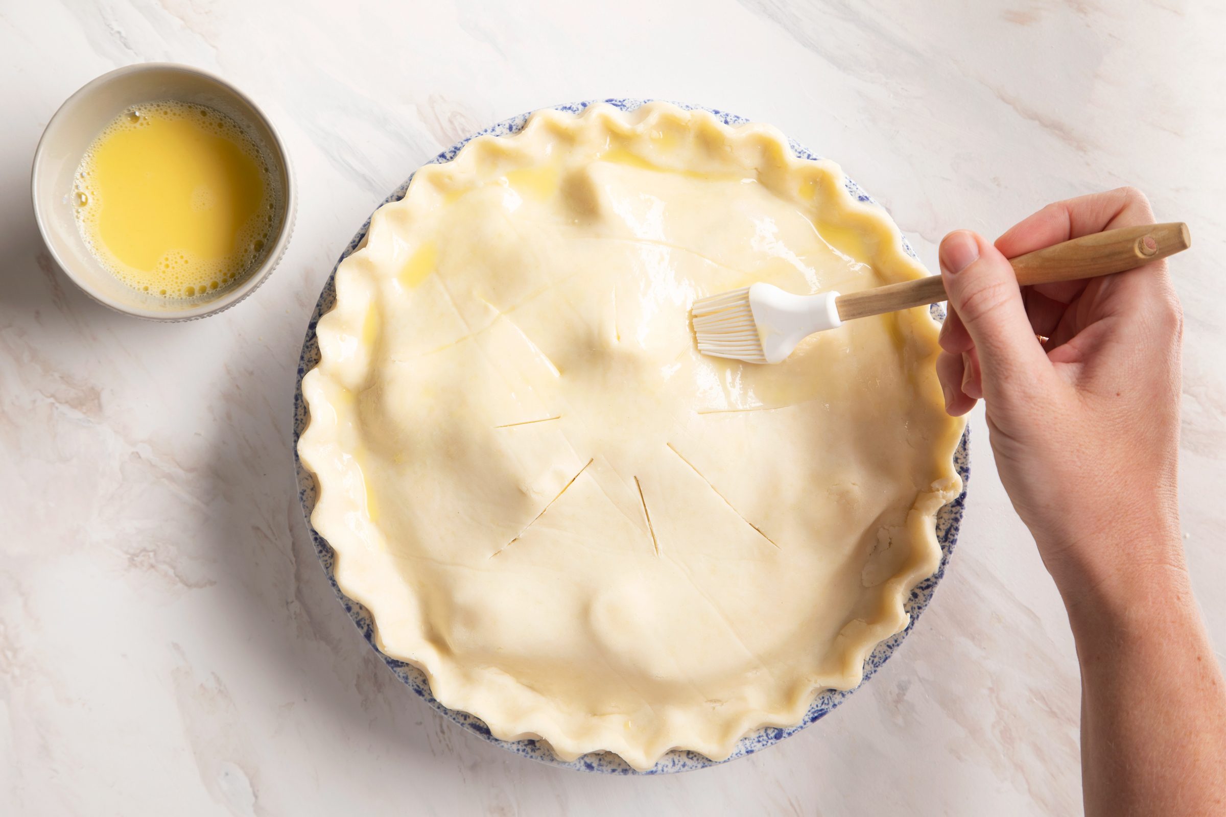 an unbaked gluten free apple pie with slits in the top and a hand brushing egg whites on the top of the crust