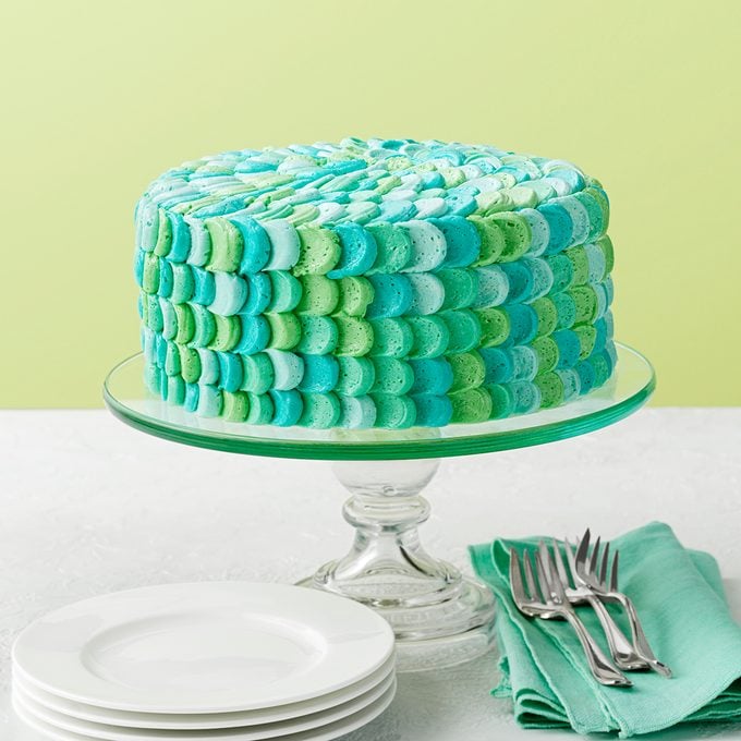 A cake with a decorative icing pattern in shades of blue and green sits on a glass cake stand. The background is light green, and nearby are stacked white plates and a folded green napkin with forks on top.