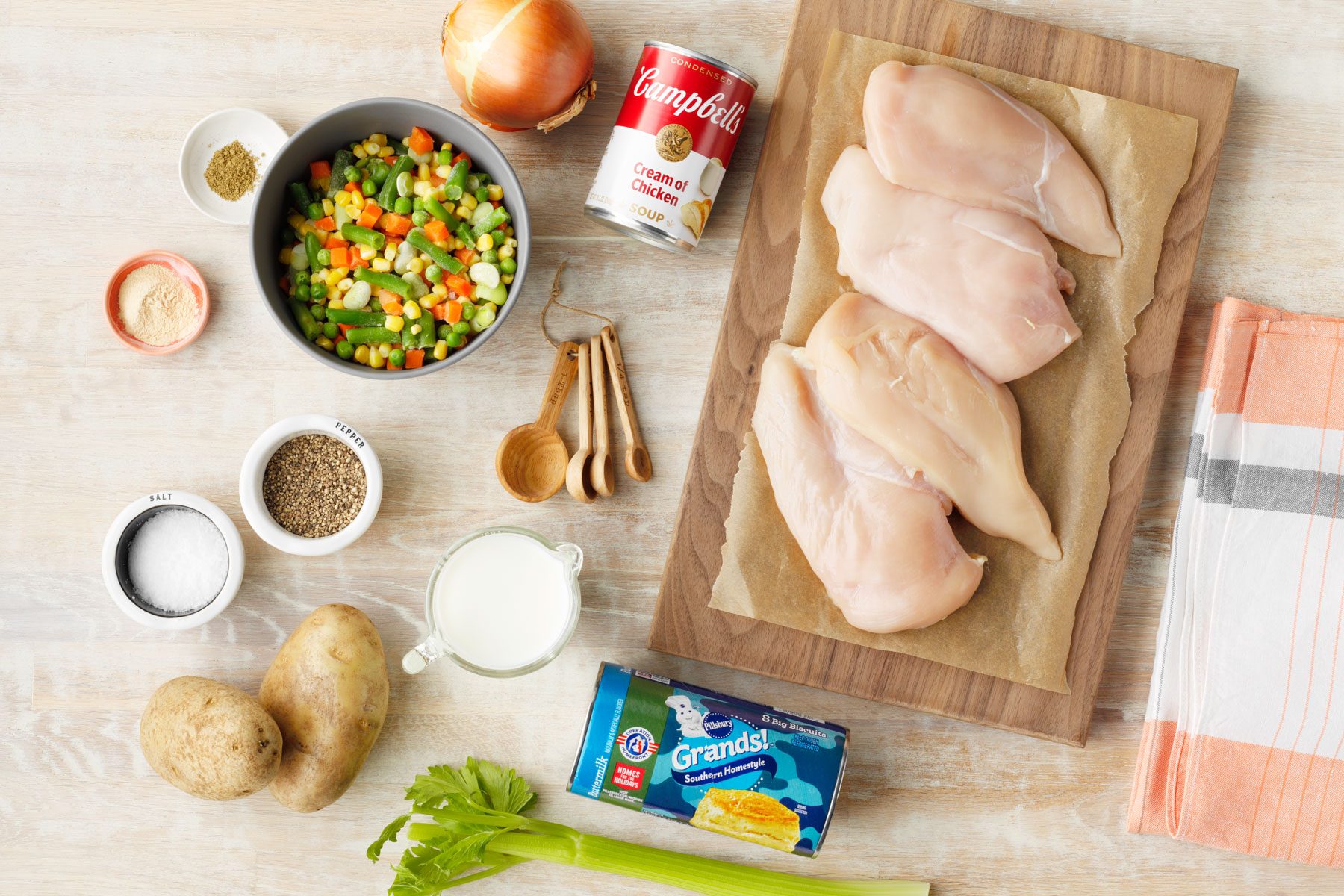 A top-down view of ingredients for a chicken pot pie laid out on a light-colored surface. Includes raw chicken breasts, mixed vegetables, a can of cream of chicken soup, potatoes, onion, seasonings, milk, celery, a cloth, and a can of biscuit dough.
