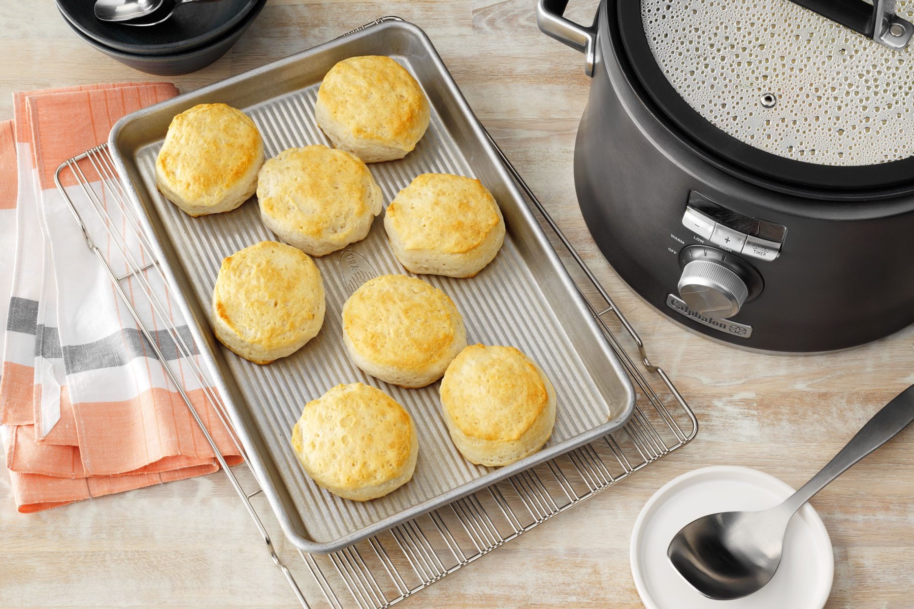 A baking tray with eight freshly baked biscuits rests on a cooling rack next to a slow cooker. There is an orange and white cloth to the left, along with an empty white bowl and a stainless steel ladle on the right. A black bowl is visible in the background.