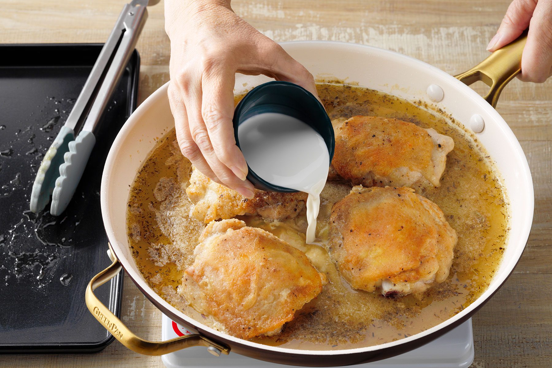 A person's hand pouring a cup of milk into a frying pan containing four pieces of crispy, golden-brown chicken. The pan has beige handles and sits on a wooden surface next to metal tongs.