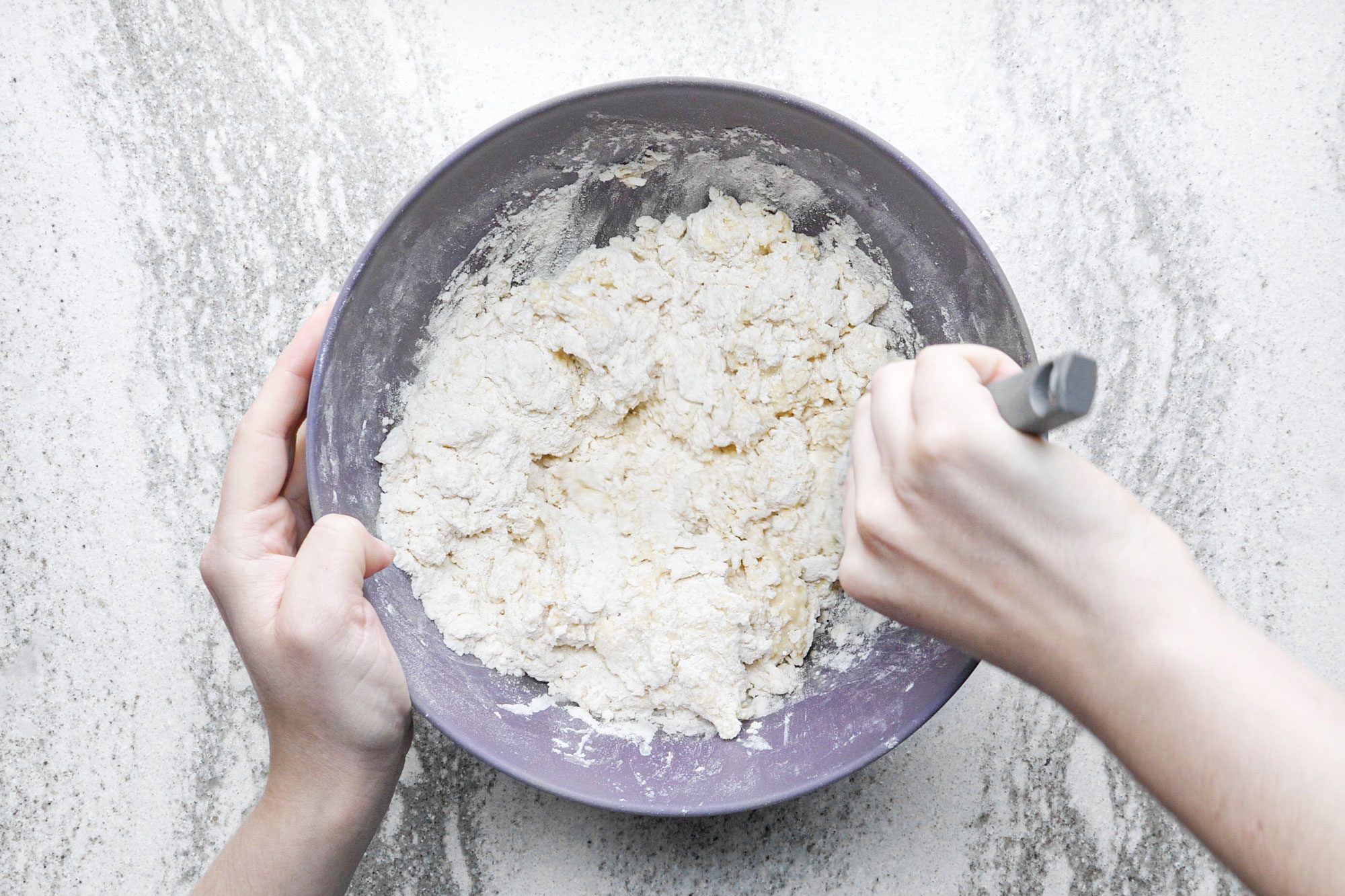 a person mixing flour and egg mixture in a large bowl on marble surface