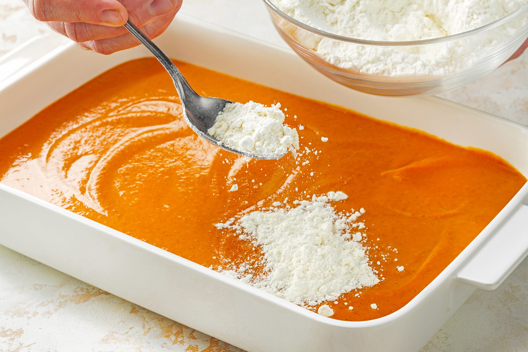 A white baking dish is filled with an orange batter. A hand is holding a spoon, sprinkling white powder from a bowl onto the batter. 