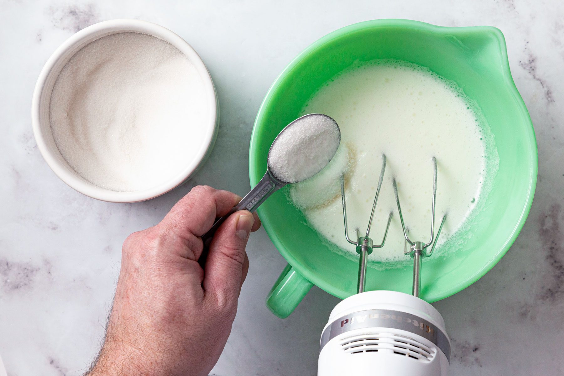 A person adding sugar in mixture in a large green bowl
