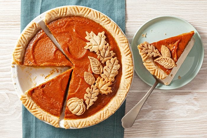 overhead shot; horizontal shot; wooden background; Pumpkin Pie in pie plate over green kitchen towel; garnishned with leaves cutout; pie piece served on small green plate with small spoon;