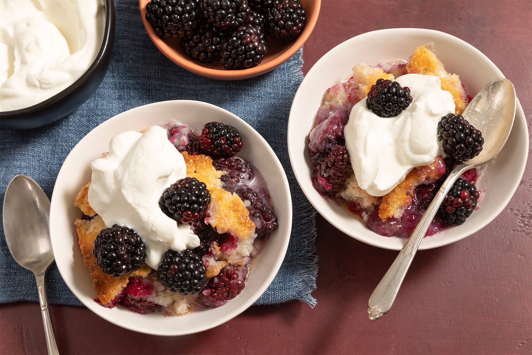 Two bowls of blackberry cobbler topped with whipped cream, accompanied by spoons, are displayed on a blue cloth. A bowl of fresh blackberries and a bowl of whipped cream are also present nearby on a red surface.