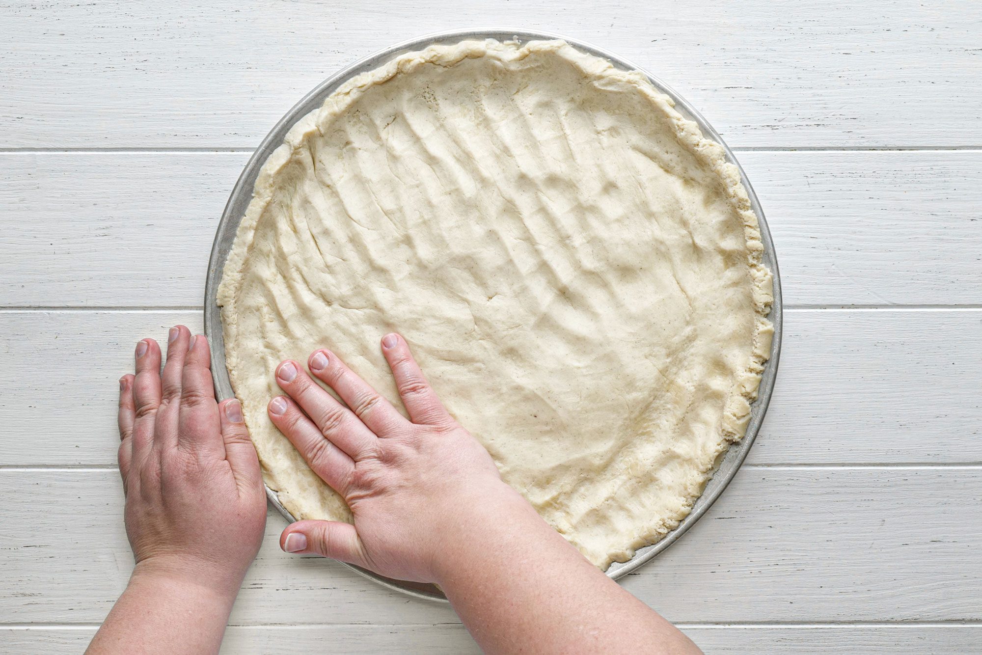 pressing the pizza crust dough on a pizza pan on white painted wooden surface