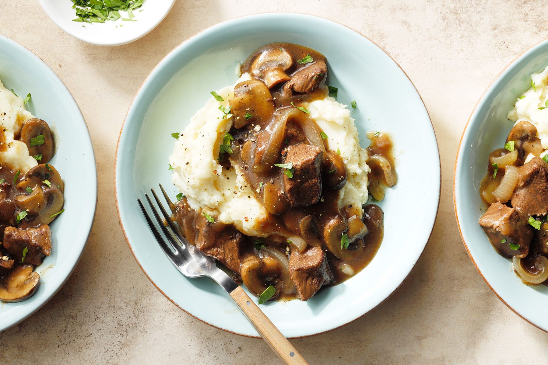 A close-up of a plate of mashed potatoes topped with beef and mushroom gravy, garnished with fresh herbs. A fork rests on the plate, and a small bowl with herbs is visible in the background. Two other similar plates are partially visible on the sides.