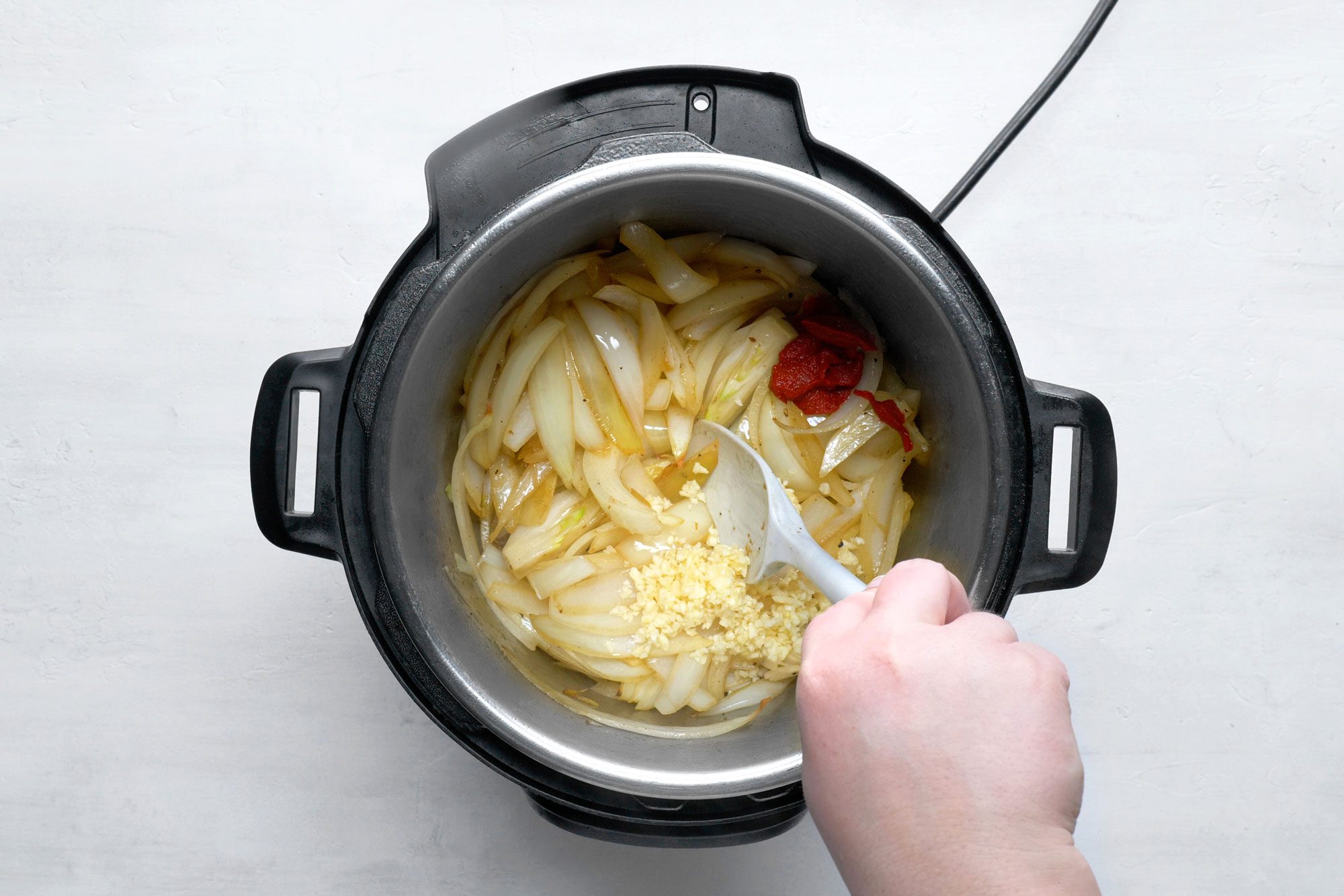 overhead shot; white background; cooking onions in electric pressure cooking using spatula added garlic and tomato paste;