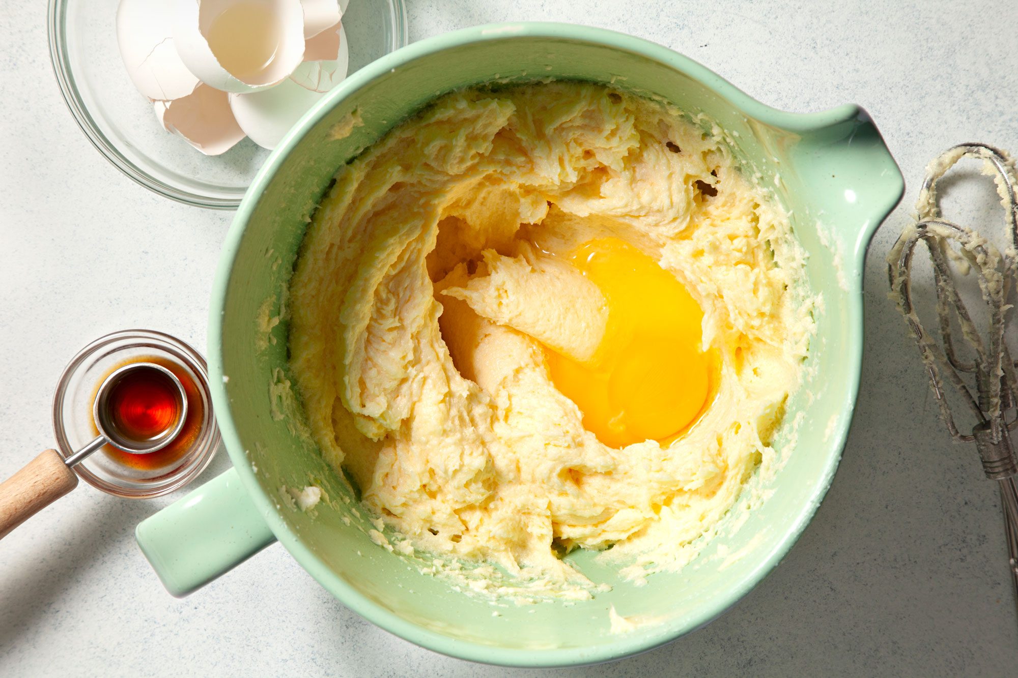 overhead shot of a light green mixing bowl contains creamy batter with eggs in the center; egg shells; vanilla extract;