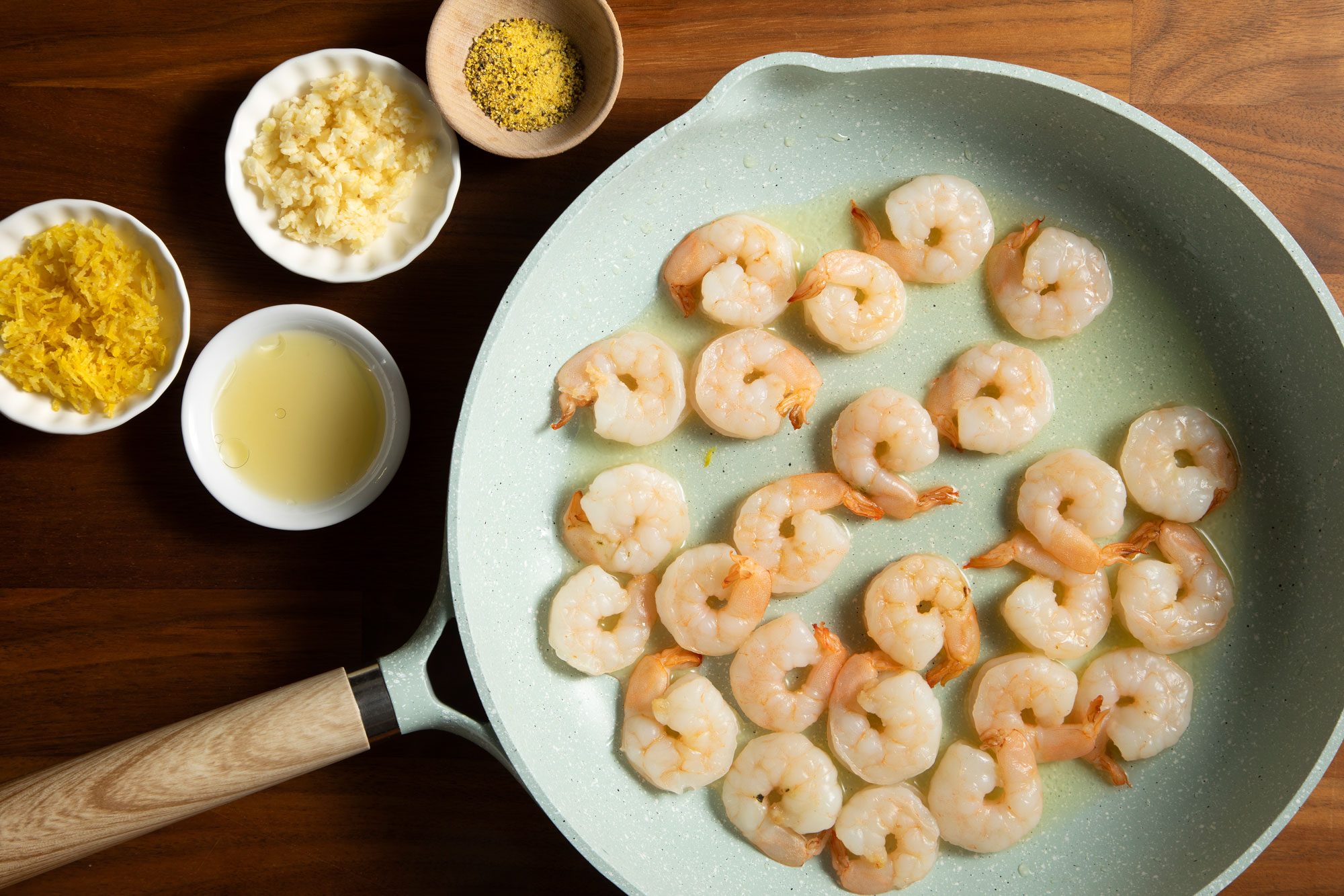 overhead shot; wooden background; cooking shrimps in large white pan; with some ingredients;