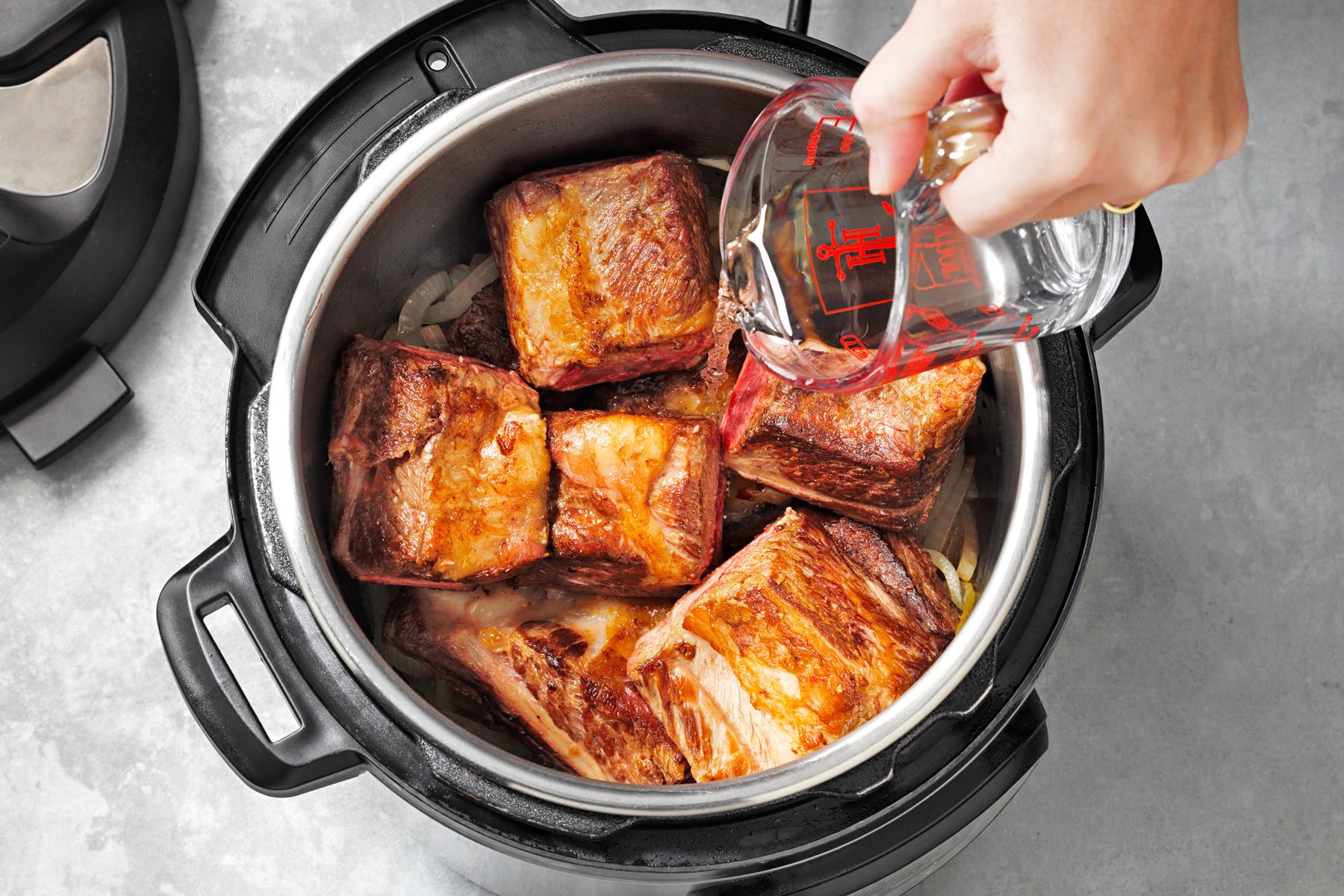 A hand pours water over beef ribs in a pressure cooker
