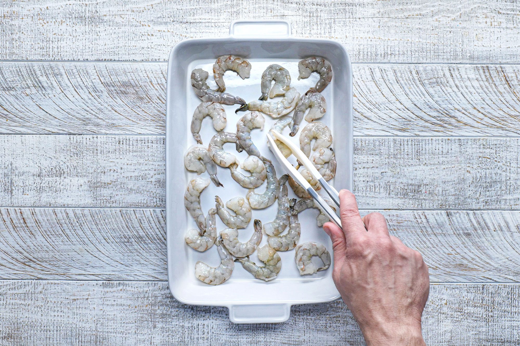 overhead shot; white wooden background; shrimps in white rectangular dish; picking shrimps using tong;