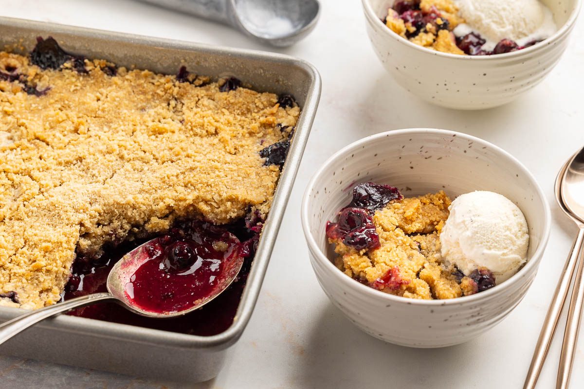 Taste of Home blueberry crisp recipe photo of blueberry crisp in baking dish, 2 bowls with dessert topped with vanilla ice cream.