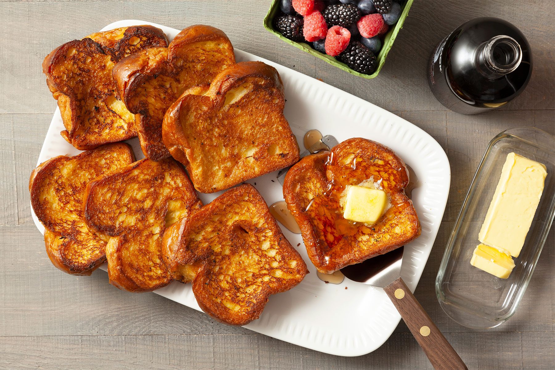 Overhead shot of Brioche French Toast; in large platter; flat spatula; syrup; butter; fresh berries; fork; light wooden background;