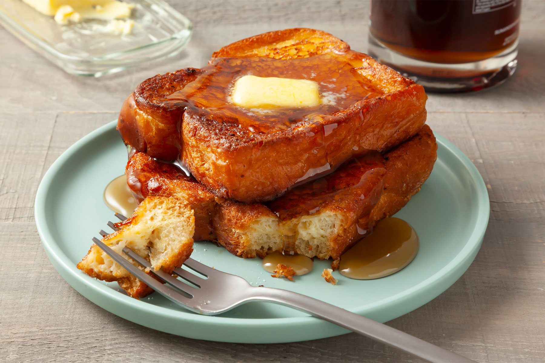 3/4 angle view shot of Brioche French Toast; served on plate top with syrup and butter; fork; light wooden background;