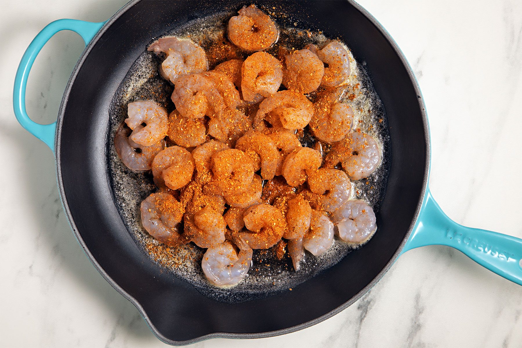 A black frying pan with a turquoise handle contains peeled shrimp being cooked. The shrimp are seasoned with a reddish-orange spice blend and are positioned on a white marble countertop.